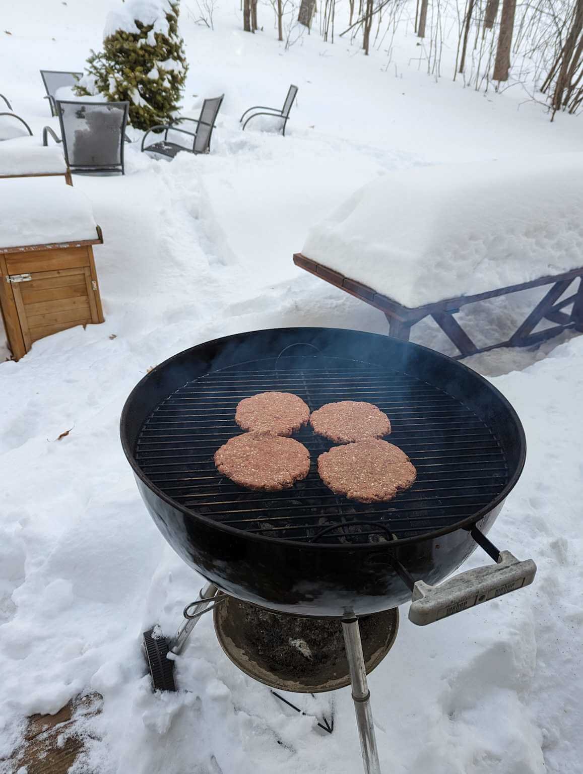 Winter grilling outdoors on a charcoal BBQ grill with snow-covered surroundings and lightly smoking hamburger patties
